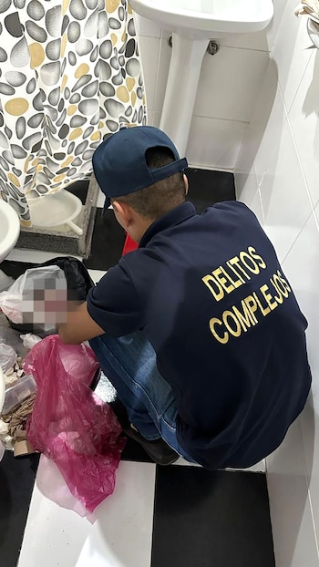 Hombre con gorra azul y uniforme de "Delitos Complejos" agachado en un baño, examinando bolsas de plástico blancas y rosadas cerca de un inodoro y un lavabo