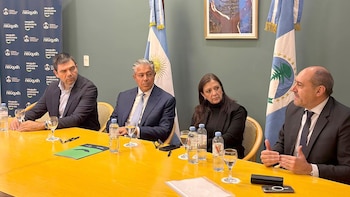 Cuatro personas, tres hombres y una mujer, sentadas en una mesa amarilla. Banderas de Argentina y Neuquén se aprecian al fondo. Hay botellas de agua y vasos en la mesa, y logos en la pared