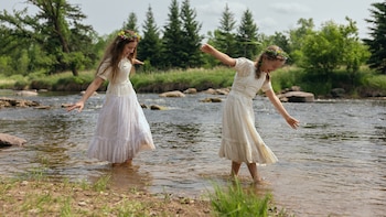 Dos niñas con vestidos largos blancos y coronas de flores, con los brazos abiertos, caminan dentro de un río con rocas, hierba y árboles
