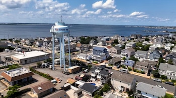 Vista aérea de Seaside Park con una torre de agua blanca prominente, numerosas casas residenciales, calles y una gran masa de agua azul al fondo bajo un cielo con nubes