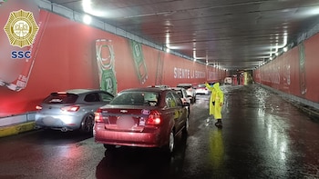 Agente de tránsito con impermeable amarillo dirige vehículos en un túnel urbano oscuro e inundado. Hay varios carros detenidos y agua en el suelo