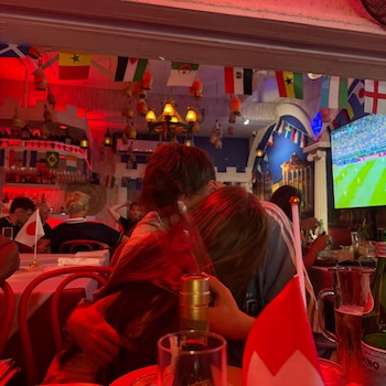 Dos personas se abrazan de espaldas en un restaurante con luz roja, banderas nacionales, un televisor mostrando fútbol y botellas en las mesas