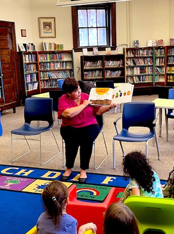Una mujer sentada lee un libro ilustrado a cuatro niños que la escuchan sentados en el suelo de una biblioteca con estanterías llenas de libros