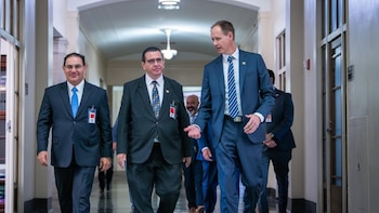 Fotografía de tres hombres en traje oscuro caminando por un pasillo iluminado. Dos de ellos llevan gafas y distintivos de identificación visibles
