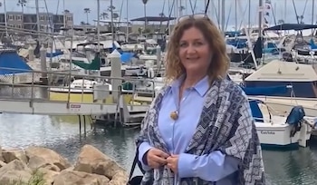 Una mujer con camisa azul y chal estampado está de pie frente a un muelle con lanchas y veleros amarrados en una marina