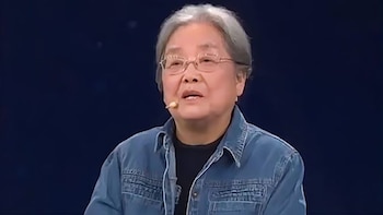 Primer plano de Cai Gao, una mujer mayor con cabello gris y gafas, vistiendo una chaqueta vaquera azul, hablando con un micrófono. Fondo oscuro