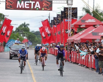 Ciclistas en bicicletas de ruta cruzan la meta frente a vallas rojas con público y un automóvil oficial detrás