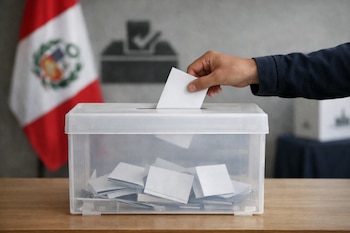 Una mano con manga azul oscuro inserta una boleta blanca en una urna electoral transparente sobre una mesa de madera clara. Al fondo, la bandera de Perú está desenfocada.
