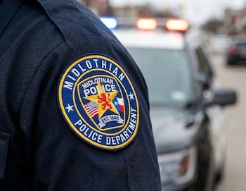 Primer plano del hombro de un oficial con el parche azul y amarillo "Midlothian Police Department". Un coche de policía con luces está borroso al fondo