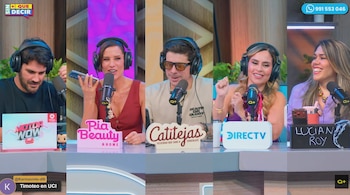 Cinco conductores de programa con auriculares frente a micrófonos en un estudio, mientras una mujer sostiene un teléfono celular en la mano
