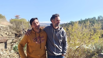 Dos hombres con sudaderas se abrazan por los hombros en un entorno rural con colinas y vegetación de fondo