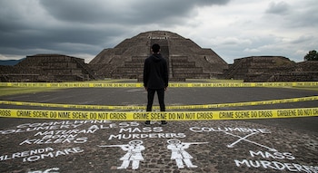 Joven de espaldas frente a la Pirámide del Sol en Teotihuacán. Cintas amarillas de "escena del crimen" rodean el área con grafitis de "Natural Born Killers" en el suelo. Cielo nublado.