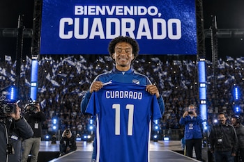 Juan Guillermo Cuadrado sostiene una camiseta azul de fútbol con el número 11 frente a una pantalla con la frase Bienvenido Cuadrado.