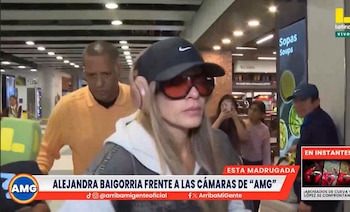 Alejandra Baigorria camina en un espacio interior, llevando gorra negra, gafas de sol rojizas y sudadera clara, acompañada por un hombre y con cámaras de fondo