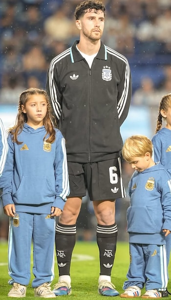 Marcos Senesi, futbolista, de pie entre dos niños pequeños vestidos con uniformes de la selección argentina en un campo de fútbol