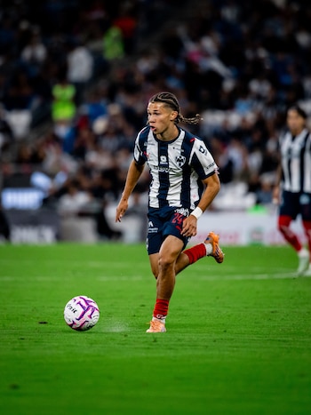 Un jugador con camiseta de franjas azules y blancas corre sobre el césped y conduce una pelota de fútbol frente a las gradas