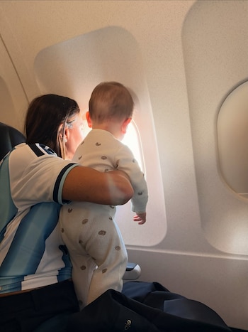 Una mujer, vestida con camiseta de Argentina, sostiene a un bebé de espaldas mirando por la ventanilla de un avión iluminada por el sol