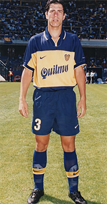 Un hombre de pie con uniforme de fútbol azul y amarillo con el número 3, la marca Quilmes y el logo de Nike, en un campo de césped con gradas llenas