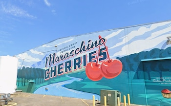 Fachada de un edificio industrial con un gran mural de cerezas rojas, texto comercial, cielo despejado, un tanque blanco y bolardos amarillos