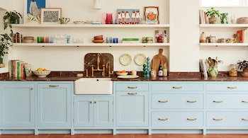 Cocina con gabinetes celestes, encimera de mármol, fregadero, grifo dorado, estantes con artículos de cocina, plantas y obras de arte.