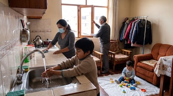 Un niño se lava las manos en el lavadero, una mujer con mascarilla limpia una superficie, un hombre abre una ventana y un bebé juega en el suelo.