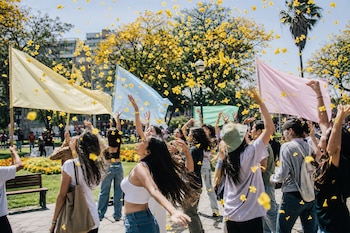 Jóvenes en un parque lanzan pétalos amarillos al aire mientras sostienen banderas de colores cerca de árboles florecidos.