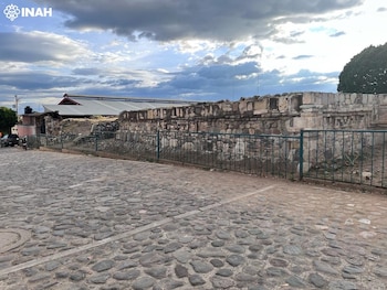 Muro de ruinas de piedra antiguo, reja de metal, calle de adoquines, cielo nublado con sol, edificios y árboles al fondo. Logo INAH
