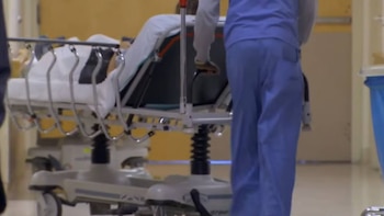 Vista trasera de un trabajador de la salud con uniforme azul empujando una camilla con un paciente cubierto en un pasillo de hospital