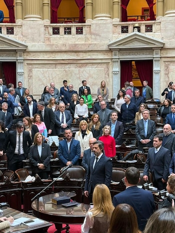 Interior de un recinto legislativo con paredes de mármol y cortinas rojas, con decenas de personas de pie y sentadas, una mesa y un micrófono