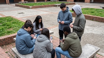 Seis adolescentes en un patio observan las pantallas de sus teléfonos móviles con logotipos de redes sociales.