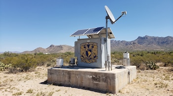 Estación sísmica gris en base de concreto, con paneles solares, antena satelital y sismógrafos. Muestra el logo de la UNAM y montañas al fondo.