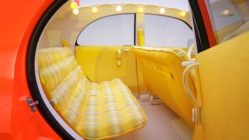 Vista del interior de un automóvil con carrocería naranja, asientos de tela amarilla a cuadros y una guirnalda de luces sobre la ventana