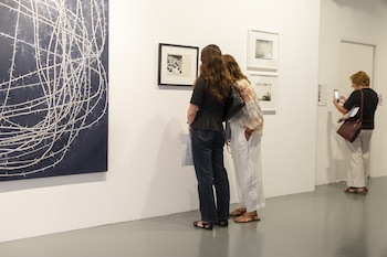 Tres mujeres observan arte en una galería moderna. Dos admiran un cuadro enmarcado, mientras otra fotografía las obras con su teléfono móvil. Las paredes son blancas