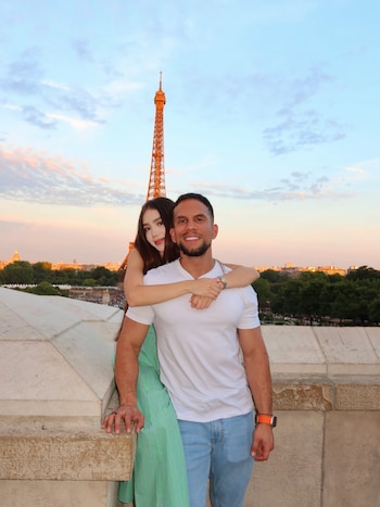 Dos personas, un hombre y una mujer, posan en una terraza con la Torre Eiffel de fondo. Cielo con nubes, vegetación y edificios