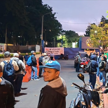Un hombre con gorra azul mira por encima del hombro, con un grupo de manifestantes y una pancarta contra el alza de combustible al fondo