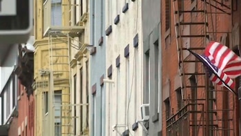 Fachada de una hilera de edificios de apartamentos en una calle urbana. Hay escaleras de incendios en varios edificios y una bandera estadounidense ondea en el último de la derecha