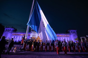Grupo de soldados con uniforme de gala sostiene una bandera de Guatemala iluminada frente a un edificio de gobierno durante la noche