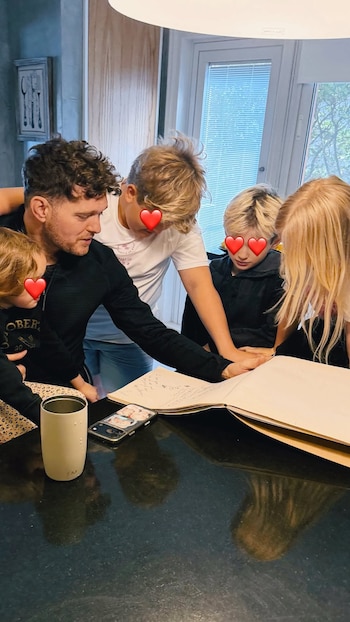 Hombre adulto y cuatro niños, tres varones y una niña, miran un libro abierto sobre una mesa oscura. Hay un vaso blanco y un teléfono celular en la mesa