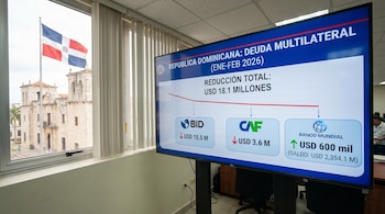Una pantalla muestra datos de deuda multilateral de República Dominicana, con la bandera dominicana y un edificio antiguo vistos a través de una ventana.
