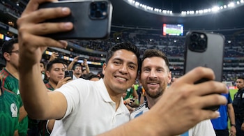 Una selfie en primera persona muestra a un aficionado latino sonriente con polo blanco junto a Lionel Messi, ambos mirando a la cámara dentro del Estadio Azteca.