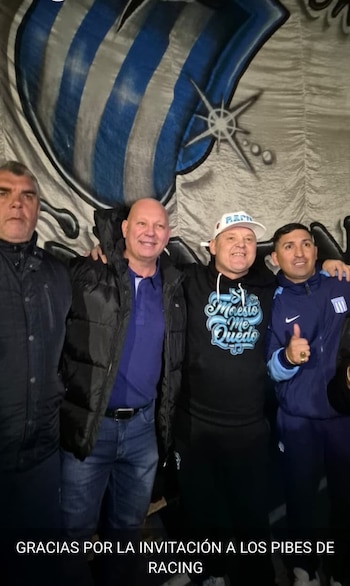 Cuatro hombres posan sonrientes delante de una bandera del club Racing. Uno lleva una gorra con la palabra Racing y una sudadera negra con texto