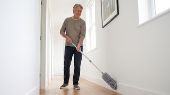 Hombre de edad media con suéter beige limpia el zócalo de un pasillo blanco con piso de madera, usando un plumero extensible gris.