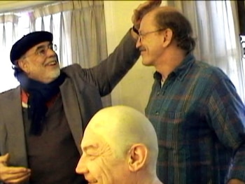 Francis Ford Coppola, con boina y bufanda, sonríe y alza la mano hacia Alex Mathews en un espacio interior