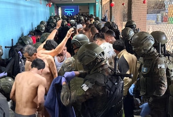 Hombres en uniforme militar y cascos, con batones, supervisan a un grupo numeroso de hombres, algunos sin camisa, en un pasillo abarrotado