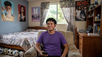 Joven con camisa morada, audífonos y celular, sonríe en sillón. Su cuarto tiene cama, escritorio, ventana, pósters de música y balones.