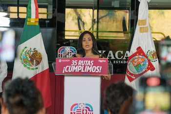Una mujer de cabello oscuro habla desde un podio frente a un autobús. Detrás de ella hay un banner rojo que dice '35 RUTAS ¡CUMPLIMOS!' y banderas