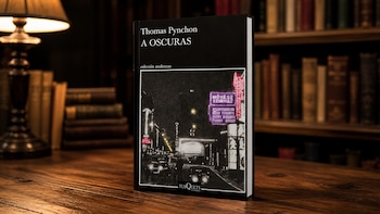 Primer plano de la portada del libro "A oscuras" de Thomas Pynchon, apoyado en una mesa de madera con una lámpara encendida y estanterías de libros antiguos en un fondo borroso
