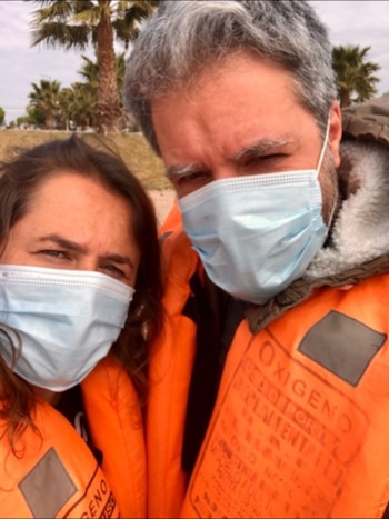 Primer plano de una mujer y un hombre con mascarillas celestes y chalecos salvavidas naranjas, con palmeras al fondo bajo un cielo nublado