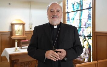 Un obispo católico vestido de traje negro y cruz pectoral permanece de pie en el interior de una capilla frente al altar y un vitral