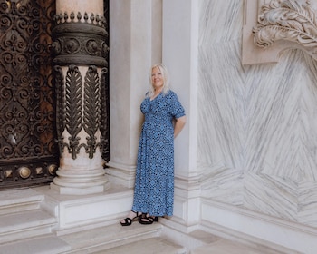 Mujer de cabello rubio con vestido azul estampado y sandalias negras, de pie junto a una columna ornamentada y una pared de mármol con diseño